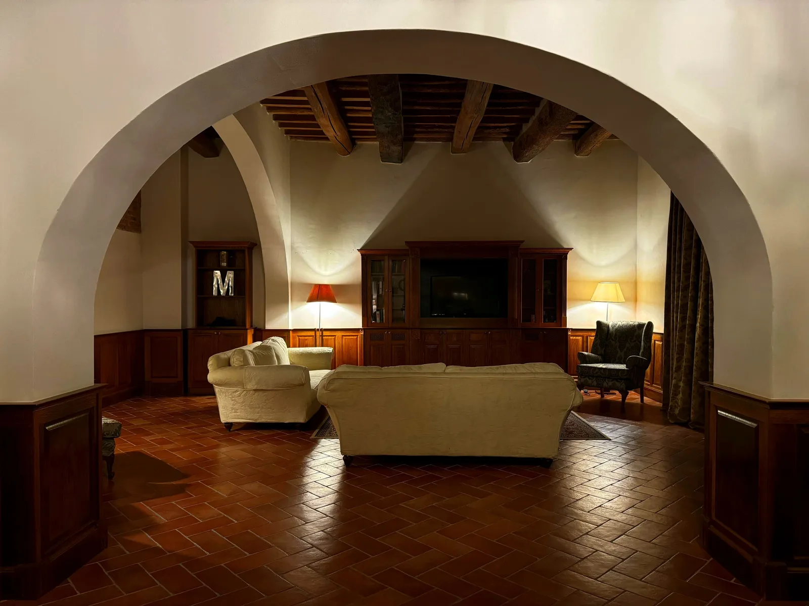 Tuscan villa living room seen through a plastered arch, with terracotta floor tiles and dark timber ceiling beams