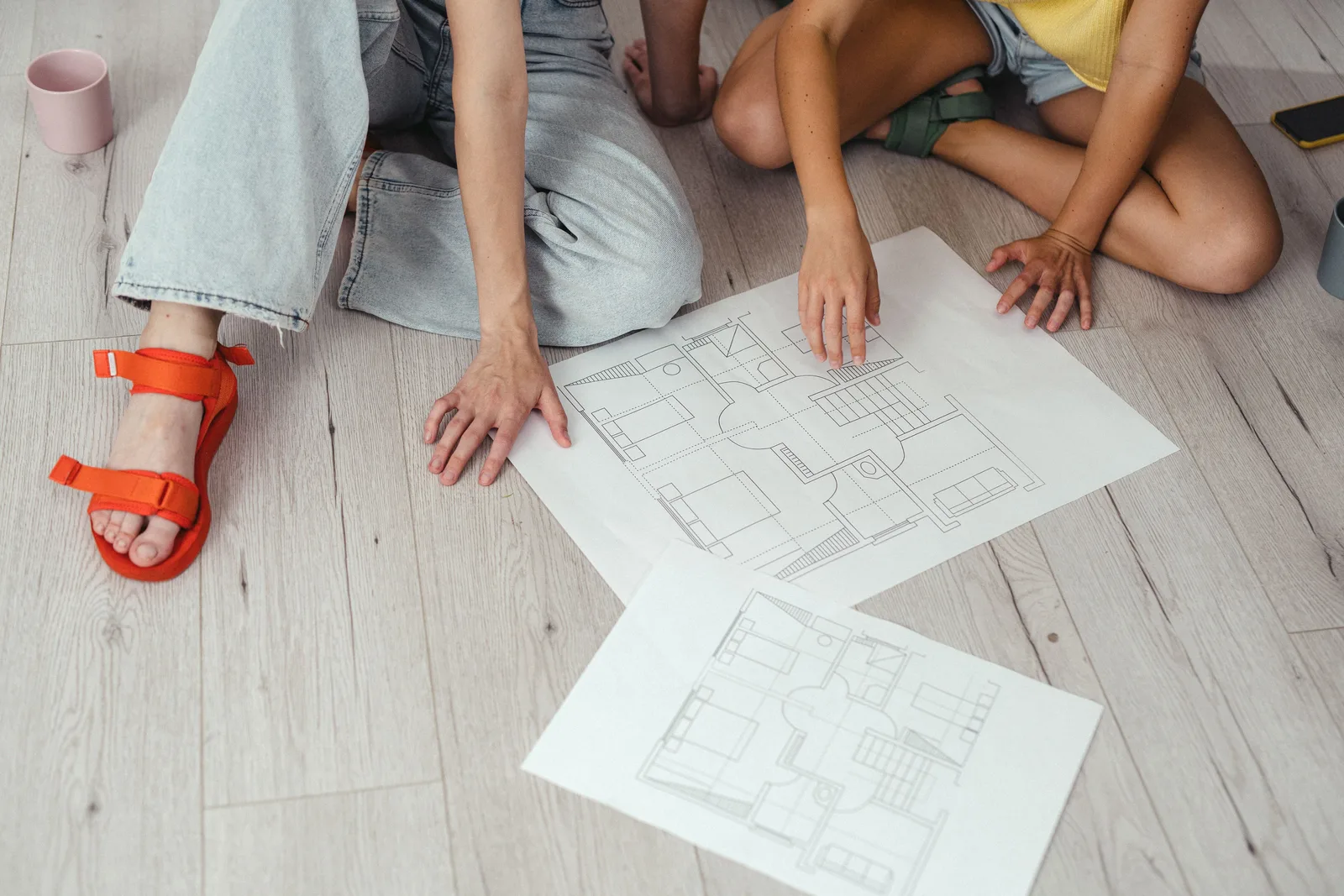 Two people on a wooden floor reviewing printed small house floor plans before building