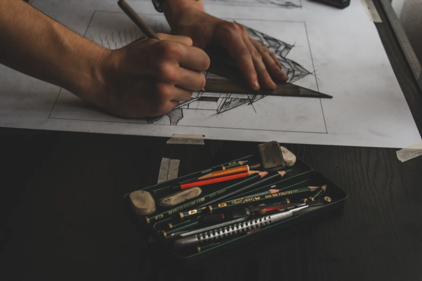 Hands drawing a pencil perspective of a house with a set square, building up linework and shading